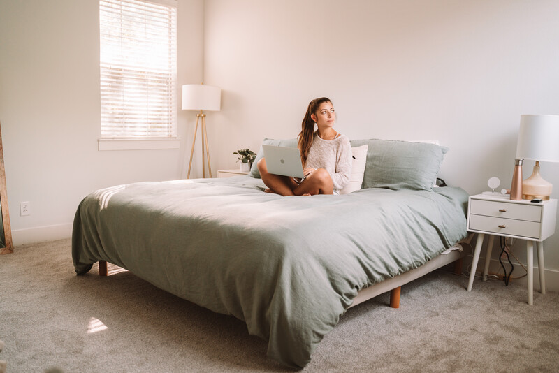 Cozy bedroom with a neatly made platform bed in sage bedding, showing a person sitting comfortably with a laptop to highlight a calm, supportive sleep and work-from-bed setup.