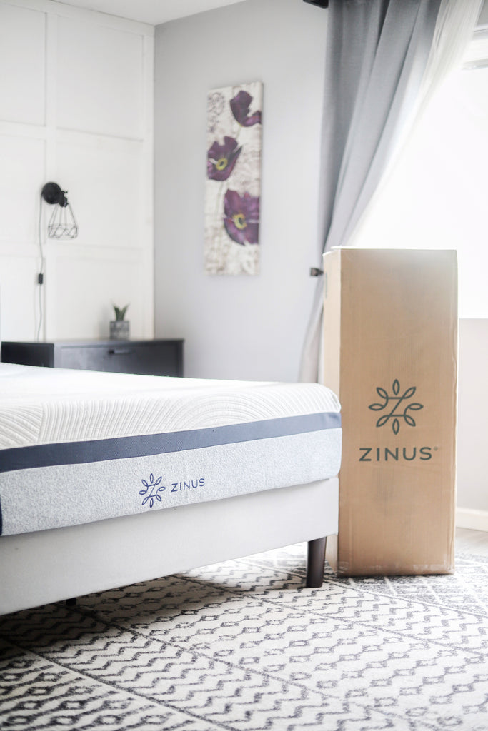 Close-up of a Zinus memory foam mattress on a light upholstered base next to its delivery box in a bright bedroom, emphasizing boxed delivery convenience and modern mattress design.