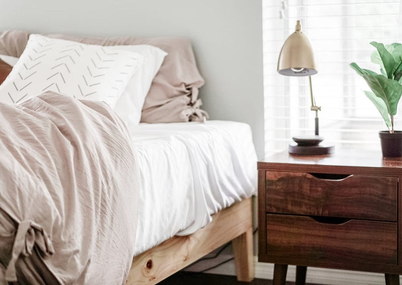 Minimal bedroom corner with a light wood bed, white bedding, soft neutral pillows, and a warm wood nightstand topped with a modern lamp and plant.