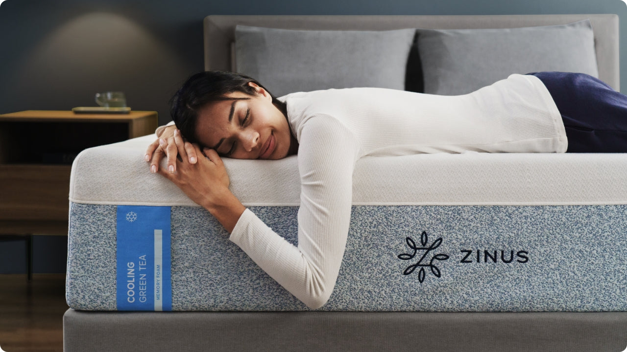 Woman relaxing against a cooling green tea memory foam mattress with a soft knit cover, showing the blue cooling tag and Zinus branding while highlighting the mattress’s refreshing feel and supportive profile in a calm, modern bedroom.