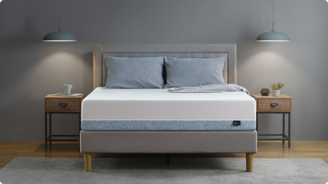 Foam mattress with a white top panel and blue-speckled side design centered in a modern bedroom, styled with a gray upholstered headboard, matching nightstands, and soft lighting to highlight its clean profile and supportive construction.