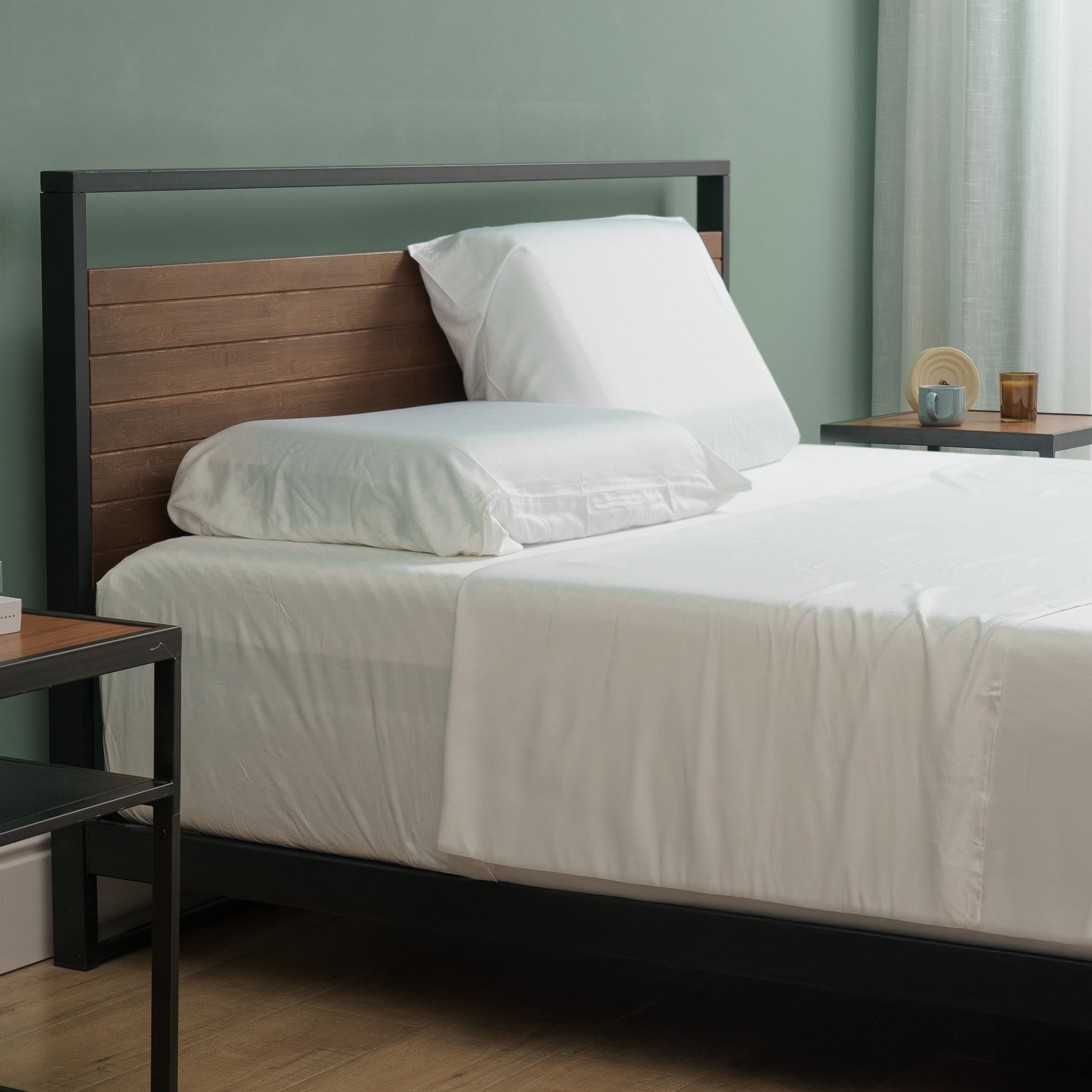 Queen bed dressed in crisp white bamboo sheet set with fitted sheet, flat sheet, and two pillowcases, styled in a modern bedroom with a black metal and wood headboard, light green walls, and nightstands holding a cup, saucer, and amber glass.