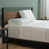 Queen bed dressed in crisp white bamboo sheet set with fitted sheet, flat sheet, and two pillowcases, styled in a modern bedroom with a black metal and wood headboard, light green walls, and nightstands holding a cup, saucer, and amber glass.