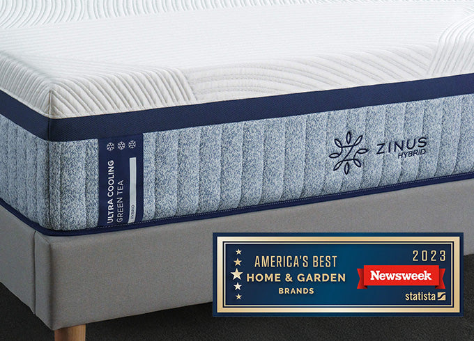 Close-up of Zinus Ultra Cooling Green Tea Hybrid mattress with textured white top and blue side panel, displayed on upholstered bed, featuring Newsweek 2023 America's Best Home & Garden Brands award badge