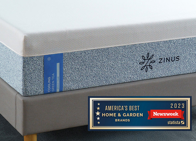 Close-up of Zinus Cooling Green Tea mattress with white top and blue patterned side panel, on upholstered platform bed, featuring Newsweek 2023 America's Best Home & Garden Brands award badge.