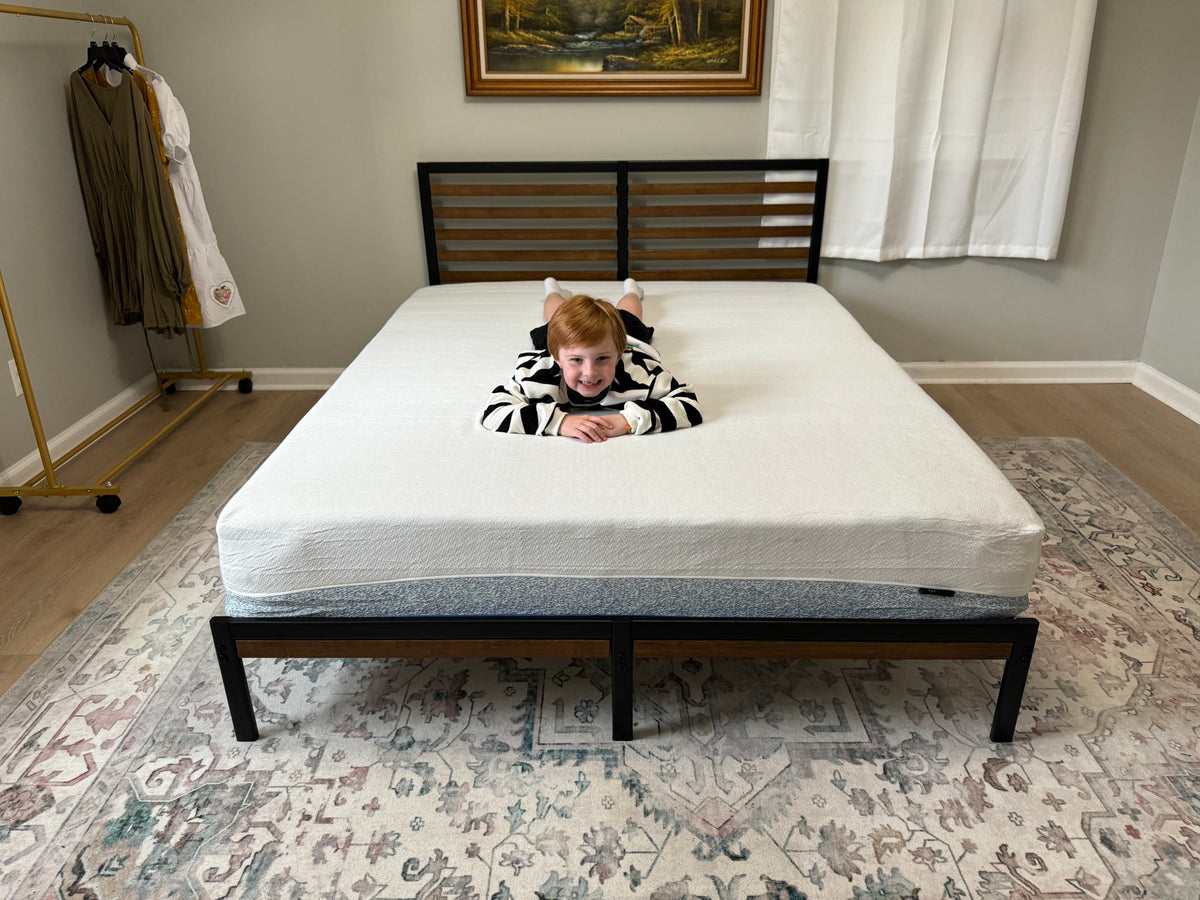 Child lying face down smiling on a Zinus mattress with white top and gray side panel, styled on a black and wood slat bed frame with patterned area rug in a simple bright bedroom, emphasizing playful comfort and supportive sleep surface