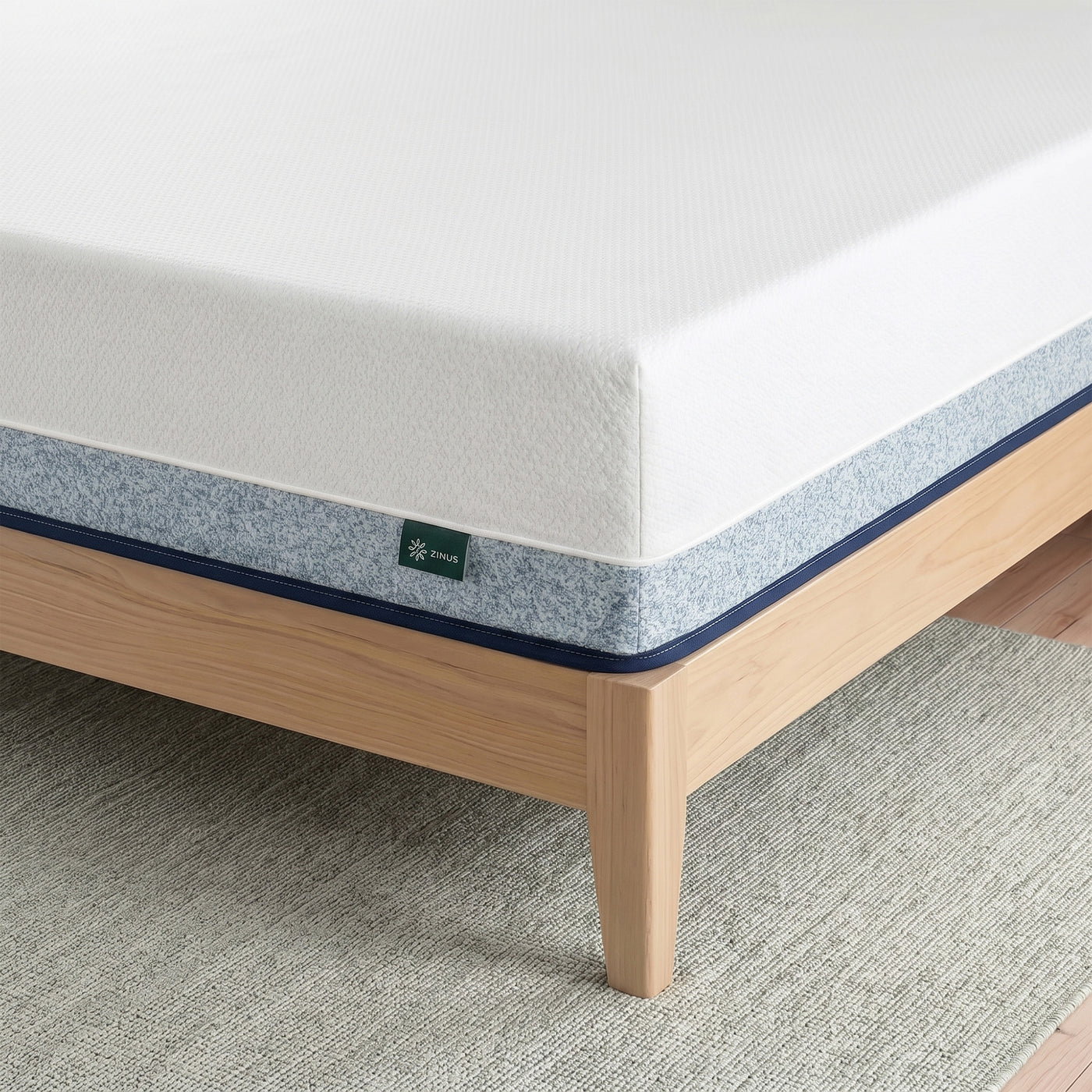 Zinus Balanced Comfort Core Hybrid Mattress, close-up corner view showing the white knit top cover, blue woven side panel with navy trim, and Zinus brand tag, set on a natural wood platform bed frame highlighting durable stitching and pocketed coil edge support.