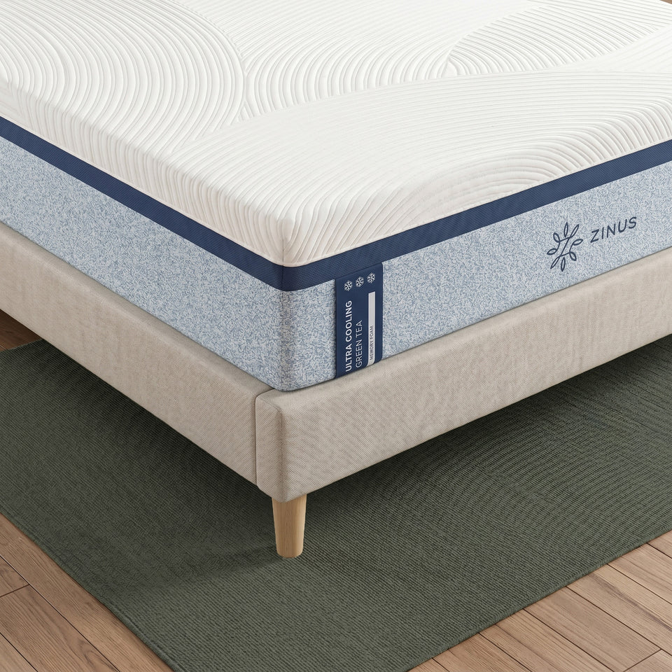 Zinus Balanced Comfort Luxe Memory Foam Mattress, close-up corner view showing the quilted white top cover with wave-pattern stitching, navy trim, and light blue woven side panel with Zinus brand tag and triple snowflake cooling icons, set on an upholstered platform bed.