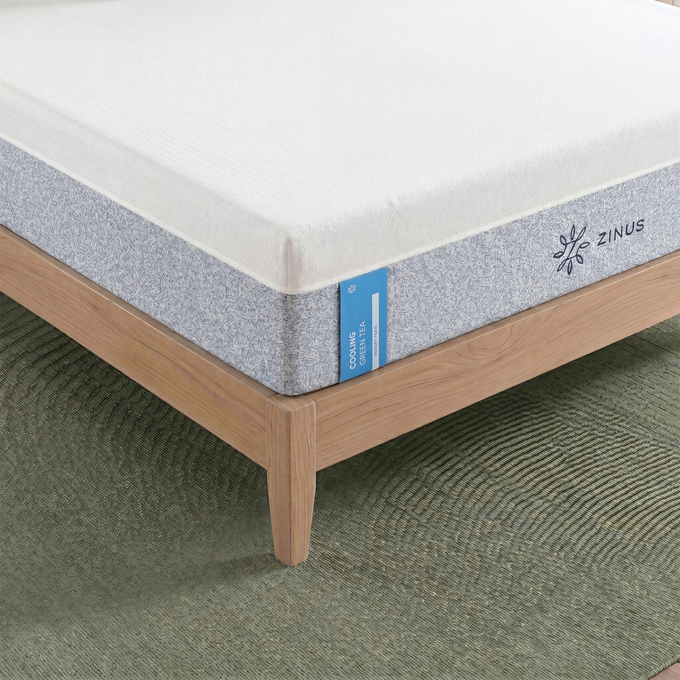 Zinus Balanced Comfort Signature Memory Foam Mattress, close-up corner view showing the white knit top cover, blue-gray woven side panel, and Zinus brand tag with a cooling icon, set on a natural wood platform bed frame with clean tailored stitching along the seam.