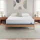 Zinus Balanced Comfort Core Hybrid Mattress fully set up on a natural wood platform bed frame in a light, airy bedroom with white and green pillows, nightstands, and lamps on either side, showing the mattress in its intended home bedroom setting.