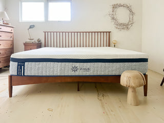 Zinus Ultra Cooling Green Tea Hybrid mattress with white top and gray patterned side panel on wooden slat bed frame, shown in a bright bedroom with wood dressers, neutral decor, and natural light, highlighting breathable cooling support in a clean modern-rustic style.