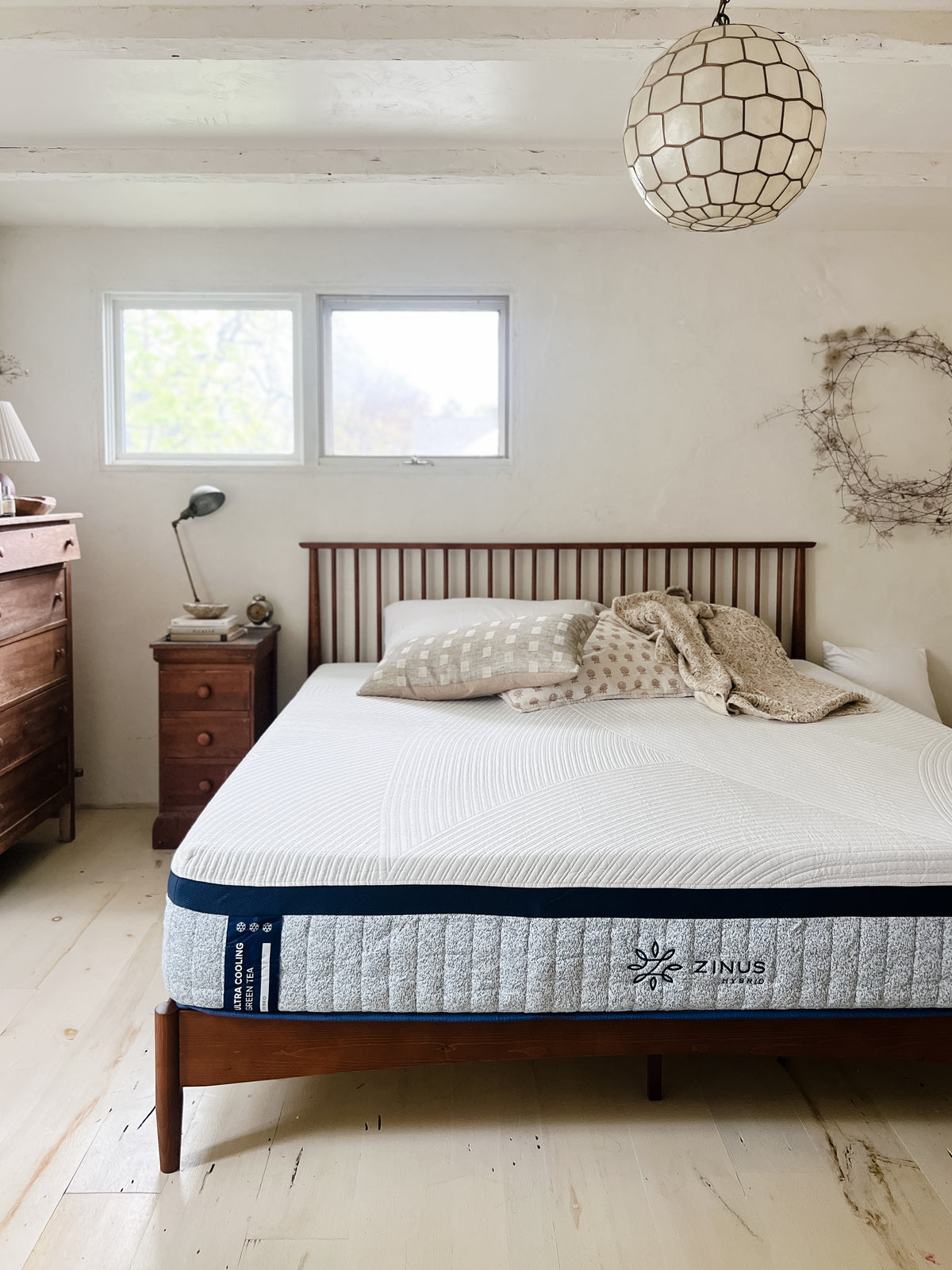 Zinus Ultra Cooling Green Tea Hybrid mattress with white top and gray side panel on wooden slat bed frame, styled with neutral pillows and blanket in a bright rustic bedroom with wood dressers, pendant light, and textured wall decor, emphasizing cooling support