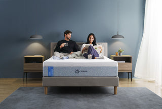 Couple sitting comfortably on Zinus mattress with white top and gray patterned side panel, relaxing with tea and reading in a modern bedroom with gray walls, pendant lamps, and wood nightstands, showcasing supportive comfort and a contemporary cozy lifestyle setting.
