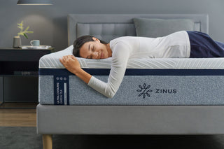 Woman in white top smiles and hugs Zinus Ultra Cooling Green Tea mattress. Visible text on mattress label reads “Ultra Cooling Green Tea Memory Foam.” Cozy modern bedroom setting emphasizes comfort and cooling mattress benefits