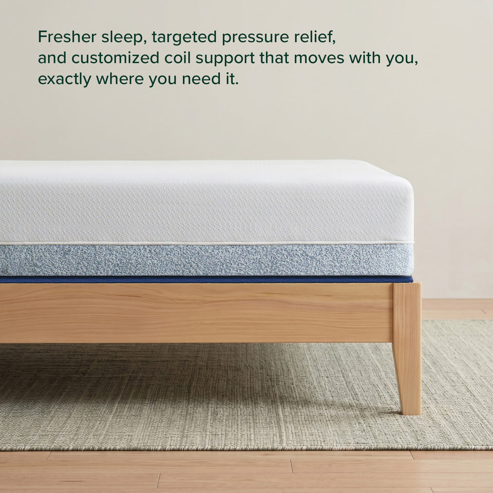 Zinus Balanced Comfort Core Hybrid Mattress, side view close-up of the white knit cover and blue woven base fabric on a natural wood platform frame, with on-image text reading: 'Fresher sleep, targeted pressure relief, and customized coil support that moves with you.