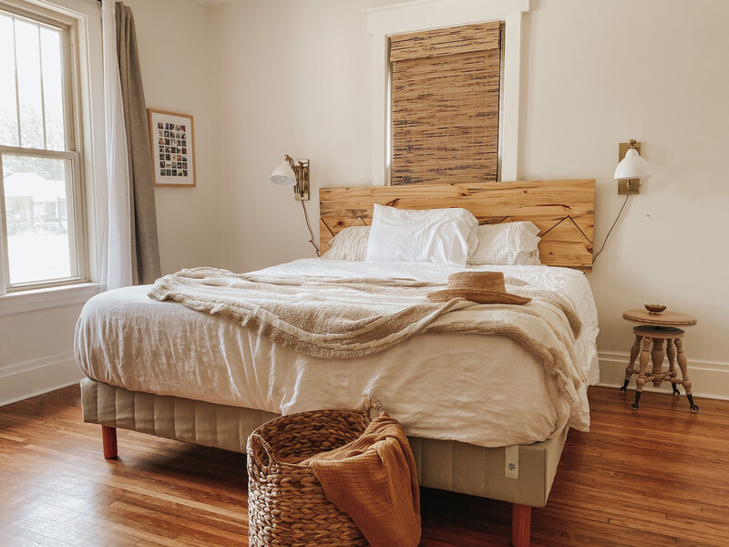 Modern bedroom with a woman sitting on a gray upholstered platform bed featuring a simple memory foam mattress and a soft pink area rug over hardwood floors.