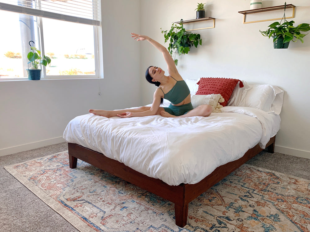 Woman stretching on a low-profile wooden platform bed with white bedding in a bright bedroom with plants and a patterned rug, highlighting a relaxed lifestyle scene and ample under-bed space.