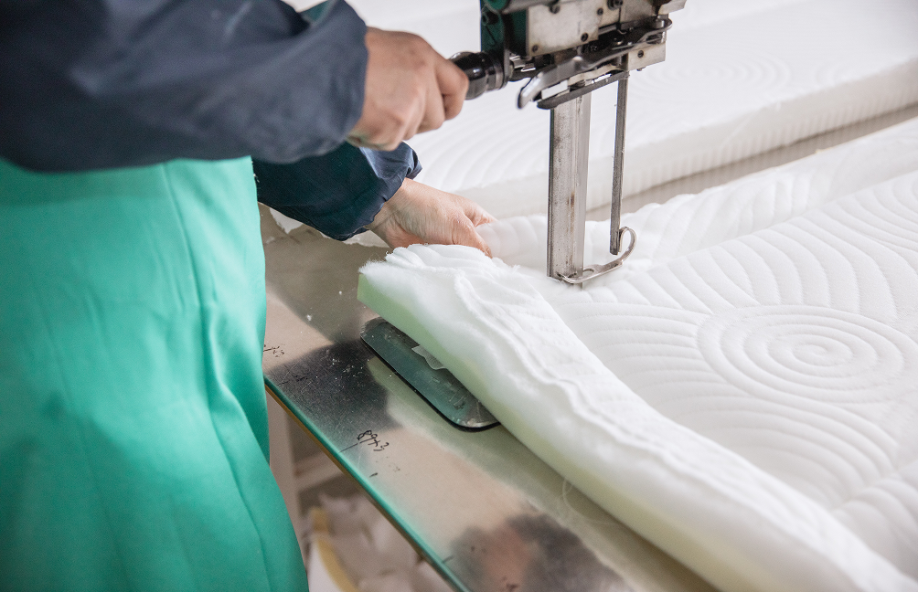 Close-up of mattress manufacturing showing a worker quilting and stitching a padded mattress cover over foam layers, highlighting careful craftsmanship, precision construction, and quality-focused production used in Zinus mattresses.
