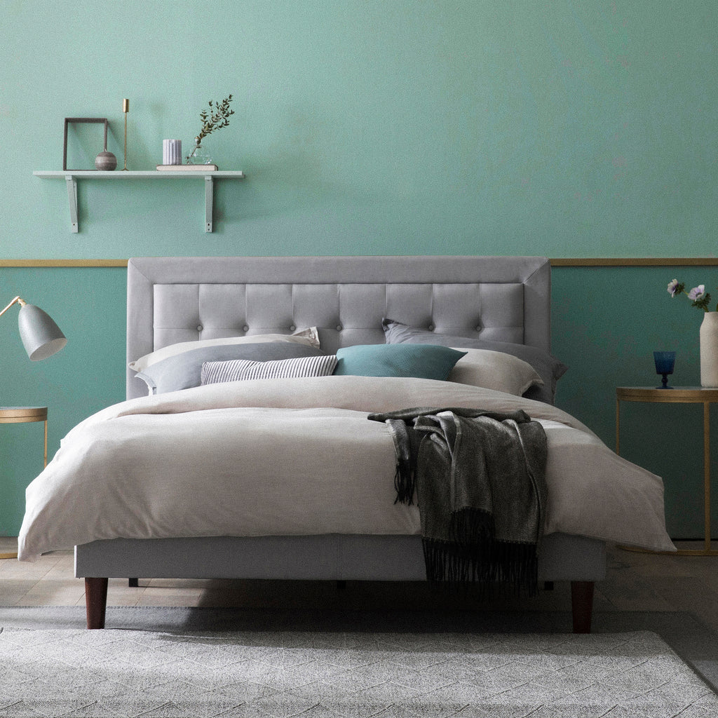 Gray upholstered platform bed with tufted headboard styled in a modern bedroom, featuring neutral bedding, accent pillows in teal and gray, and a dark throw blanket; room includes a round side table with lamp, chair, and decorative shelf against a two-tone green wall.
