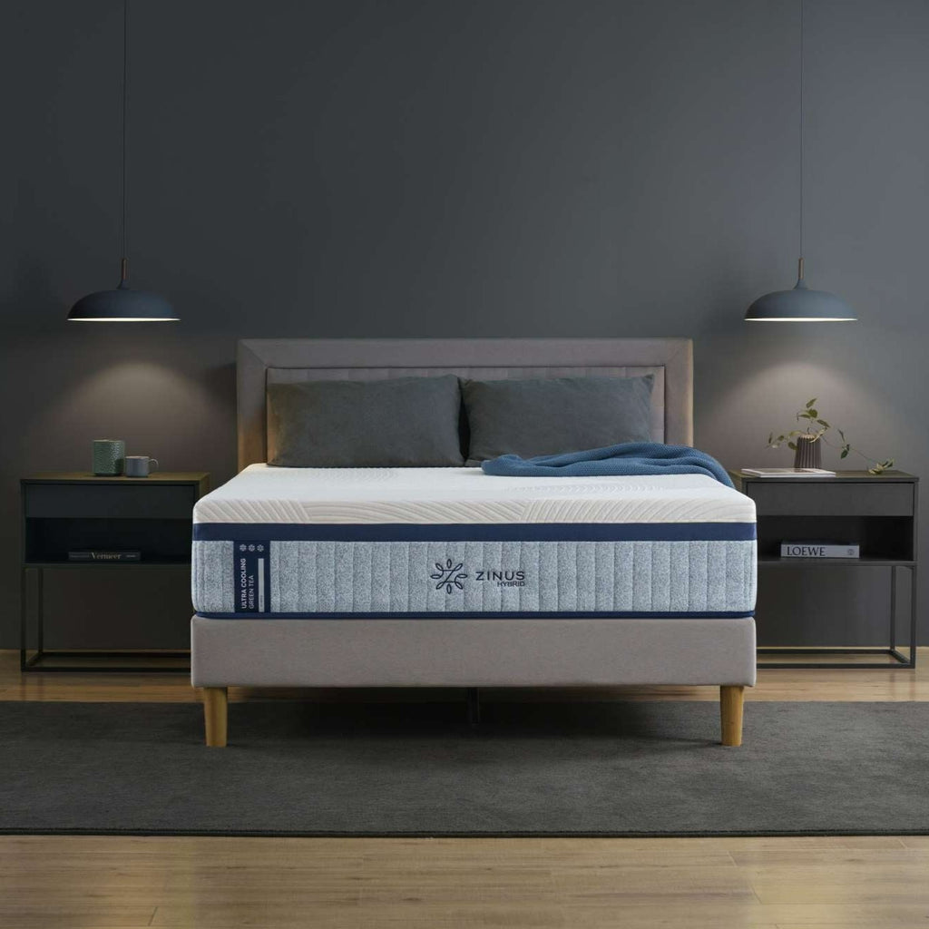 Zinus Ultra Cooling Green Tea Hybrid Mattress on upholstered platform bed frame in dark moody bedroom, showing quilted white cover with snowflake icons and ribbed gray side panel featuring Zinus logo, styled with charcoal pillows and blue knit throw between wood-topped nightstands.